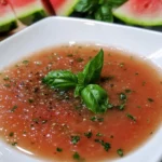 Bowl of vibrant watermelon gazpacho garnished with basil leaves and diced vegetables, served cold on a rustic wooden table
